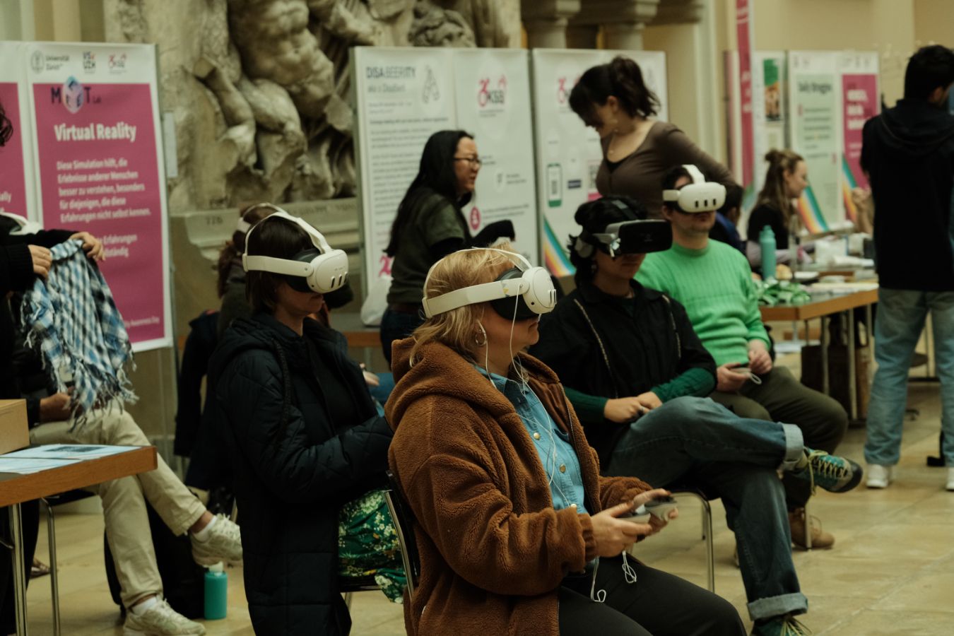 VR-Simulation: Mehrere sitzende Personen tragen Virtual-Reality-Brillen und nutzen Controller, während Betreuende und Infostände im Hintergrund zu sehen sind.